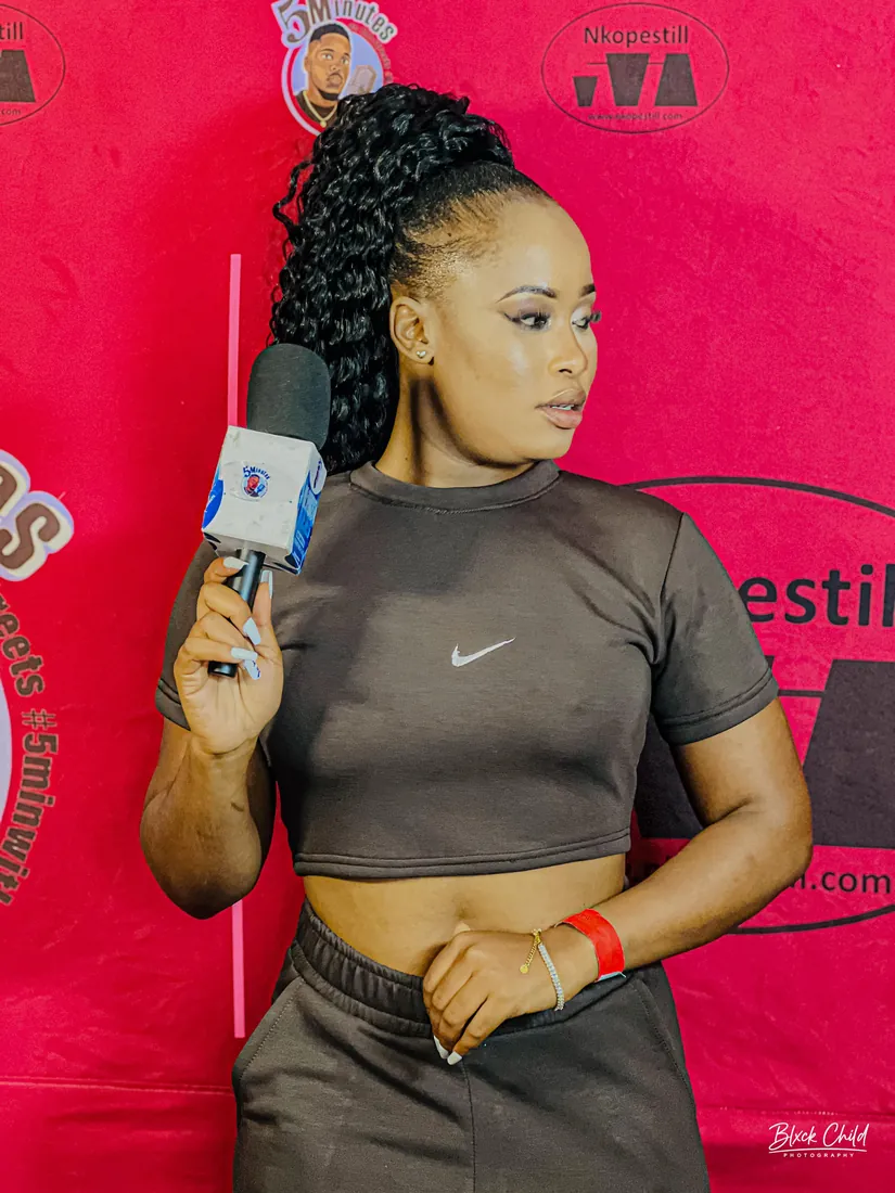 Nthabi hosting a brand event in front of a sponsor wall, branded microphone in hand